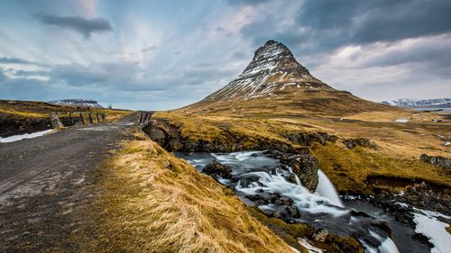 Empty dirt road by stream against mountain during winter