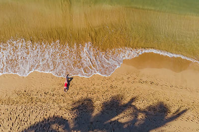 Woman lying on the beautiful sandy beach touching ocean waves with her feet, drone aerial view.