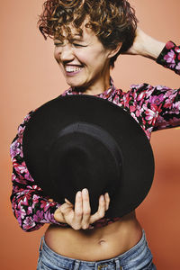 Fashionable happy female model holding a stylish black hat in colorful crop top with floral print standing against orange background with closed eyes