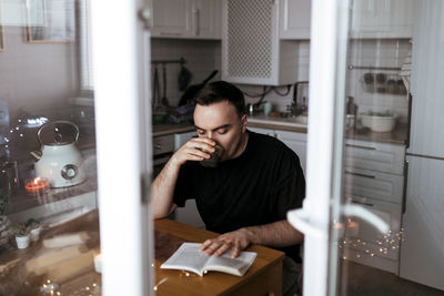 Young man drinking tea and reading a book in his modern light kitchen