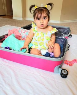 Portrait of cute baby girl sitting in suitcase on floor at home