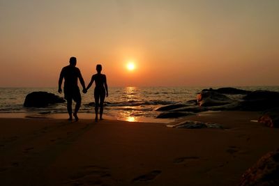 Rear view of silhouette man and woman holding hands while walking on shore during sunset
