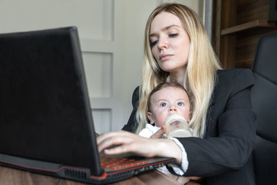 Young business woman feeds a baby from bottle in office, combining it with work on laptop.
