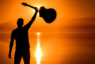 Silhouette man holding guitar while standing against orange sky during sunset