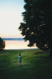 Girl standing on grassy field against tree during sunset