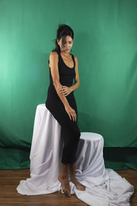 Portrait of adult model sitting on wooden chair against dark green background. 