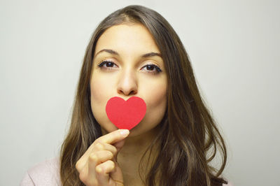 Portrait of beautiful young woman holding heart shape paper against gray background