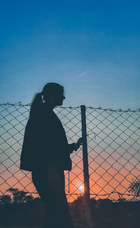 Silhouette woman standing by fence against sky during sunset