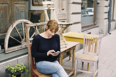 Woman using mobile phone while sitting on chair against store