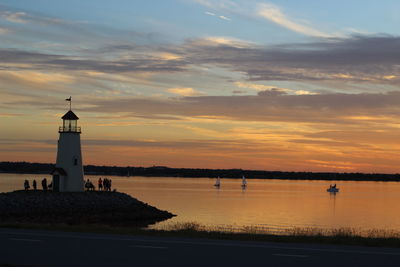 View of tourists visiting lighthouse by lake against cloudy sky during sunset