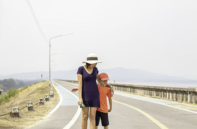 Mother with arm around son walking on road against sky