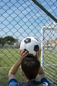 Rear view of boy holding soccer ball while standing by fence at dugout