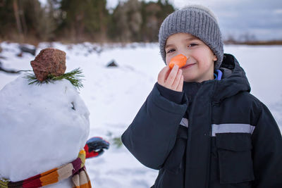 Boy in hat bites carrot, which was the nose of snowman, smiling. winter family weekend, lifestyle