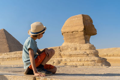 The boy in hat sits in front of the sphinx and looks at it
