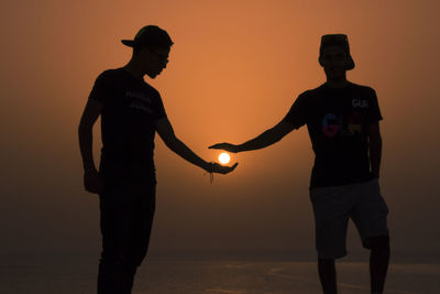 Silhouette friends holding sun while standing at beach against orange sky during sunset