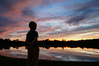 Silhouette boy standing at lakeshore against sky during sunset