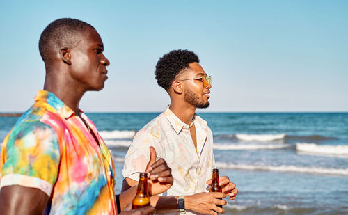 Side view of young stylish african american male friends chatting and admiring waving ocean while standing on beach with bottles of beer during summer vacation