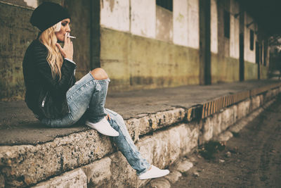 Full length side view of young woman smoking cigarette while sitting against building