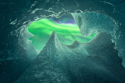 From below ice peaks surrounding hole in cave ceiling against glowing green polar lights on winter day in vatnajokull national park in iceland