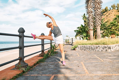 Young beautiful sportive woman training over seaside promenade, stretching legs before jogging. 