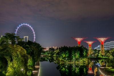 Illuminated ferris wheel by river against sky at night