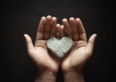 Cropped image of hand holding heart shape stone against black background