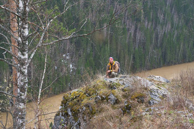 A tourist in a brown jacket and a bright camouflage bandana sits on a high bank of a mountain river. 