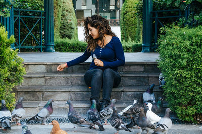 Full length of young woman feeding pigeons while sitting on steps