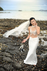Full length portrait of smiling bride holding high heels while standing at beach