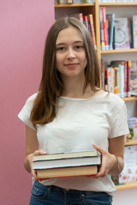 Portrait of smiling teenage girl holding book while standing in library