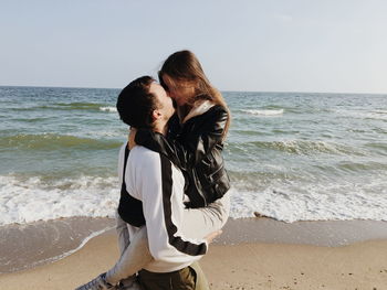Boy kissing woman while carrying at beach against sky