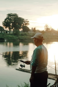 Rear view of man standing by lake against sky during sunset