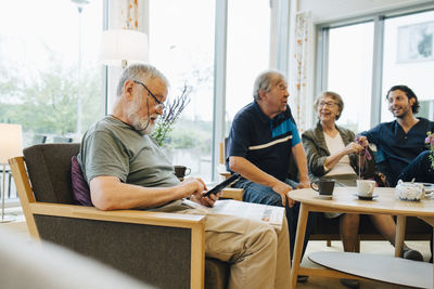 Senior man using smart phone while sitting with newspaper on armchair by friends and caretaker at elderly nursing home