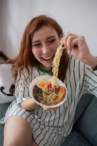 From above cheerful redhead female laughing and picking noodles from bowl of tasty ramen while sitting on couch at home