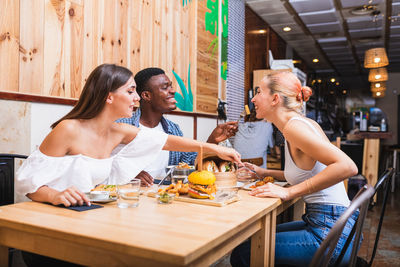 Positive young multiracial friends eating tasty dishes and burgers while gathering in modern restaurant