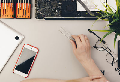 Cropped image of woman holding tweezers with mobile phone and eyeglasses on desk