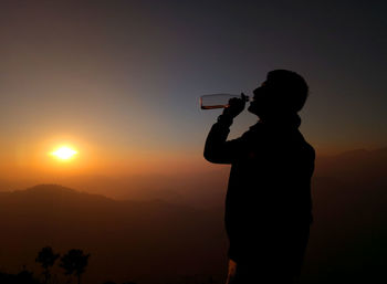 Silhouette man drinking water from bottle against sky at sunset