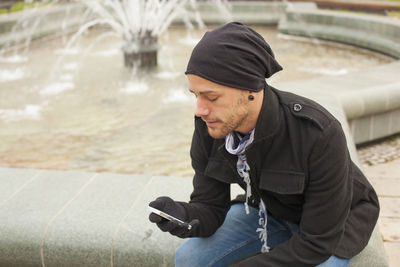 Young man using mobile phone while sitting against water fountain in city