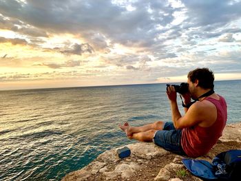 Man photographing while sitting on sea against sky during sunset