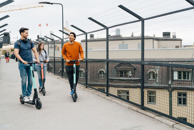 Full length of friends using push scooters on bridge in city against sky