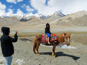 Teenage girl photographing sister sitting on horse against mountains