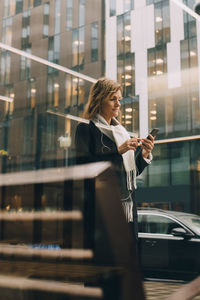 Side view of businesswoman using mobile phone while walking on sidewalk against building in city