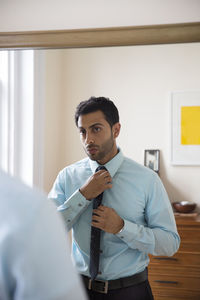 Man adjusting necktie while standing in front of mirror at home