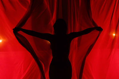 Silhouette of unrecognizable female opening light red curtains with spread arms while standing in murk room at dark night time