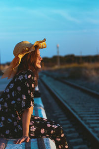 Side view of smiling young woman wearing hat sitting on railroad station platform against sky during sunset