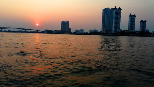 Modern buildings by sea against sky during sunset