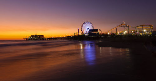 Illuminated amusement park by sea against sky at sunset