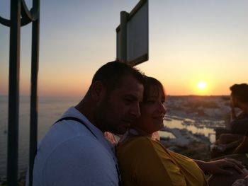 Couple looking away while standing by railing against sky during sunset