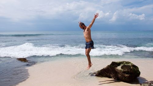 Cheerful shirtless man jumping on shore at beach against sky