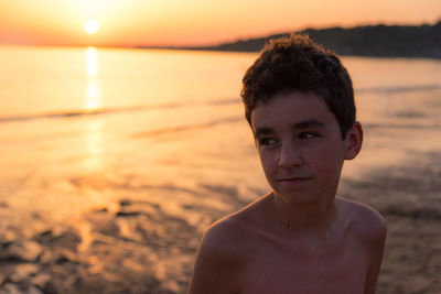 Shirtless boy looking away while standing at beach against sky during sunset